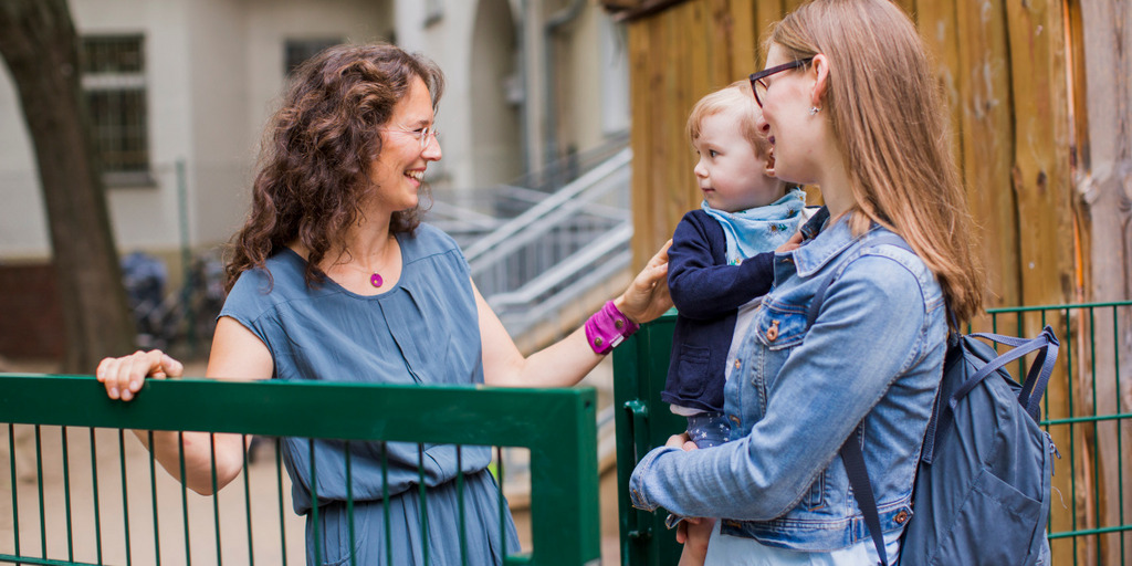 Zwei lächelnde Frauen stehen am Zaun einer Kita. Die rechte lehnt am Zahn, die linke steht ihr gegenüber und hält ein Kleinkind im Arm.