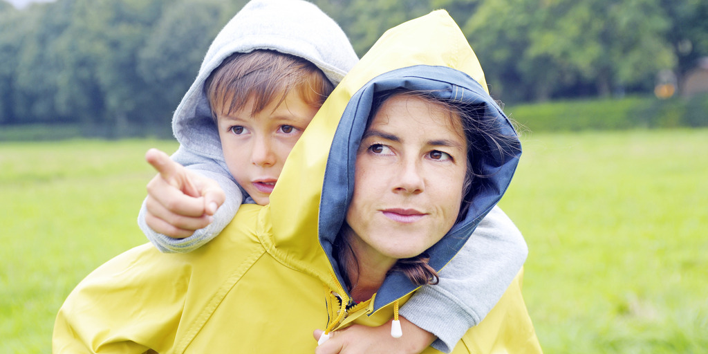 Eine junge Mutter trägt ihren kleinen Sohn über eine Wiese Huckepack. Beide tragen Regenjacken. Der Junge deutet in die Ferne, die Mutter blickt in die Richtung, in die er zeigt.