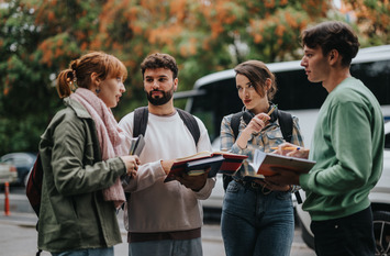 Das Bild zeigt vier junge Erwachsene im Gespräch – vermutlich Studierende – im Freien vor einem Bus. Sie halten Bücher und Notizblöcke in den Händen und wirken konzentriert und engagiert.