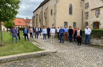 Eine Gruppe steht vor einem alten Gebäude, auf einem steinernen Weg. Links und rechts befindet sich Rasenfläche.