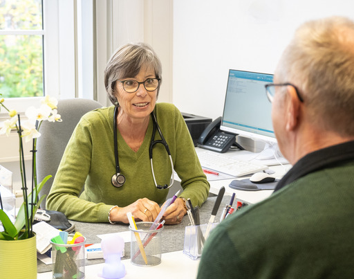 Das Bild zeigt eine freundliche Szene in einer Arztpraxis: Eine Ärztin im Gespräch mit einem älteren Patienten. Beide sitzen sich an einem Schreibtisch gegenüber. Die Ärztin trägt ein Stethoskop um den Hals und hört aufmerksam zu. Im Hintergrund ist ein Computerbildschirm zu sehen, auf dem medizinische Daten angezeigt werden.