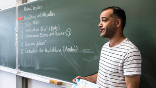 Ein Lehrer steht zu seine Teilnehmenden gerichtet vor einer grünen Tafel.