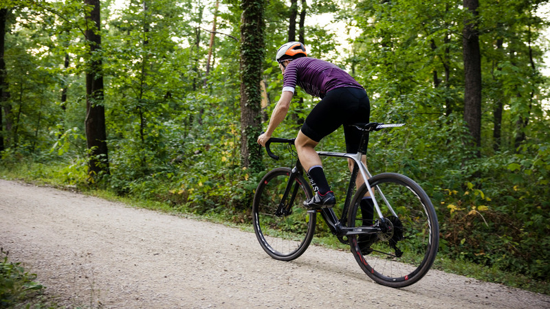 Das Bild zeigt einen Radfahrer, der auf einem Schotterweg durch einen dichten, grünen Wald fährt. Der Fahrer trägt eine sportliche Radlerkleidung, einen lila Radtrikot und einen Helm.