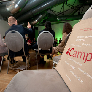 im Vordergrund: eine Tasche mit dem Aufdruck "Camp Q", im Hintergrund: ein Stuhlkreis mit Personen