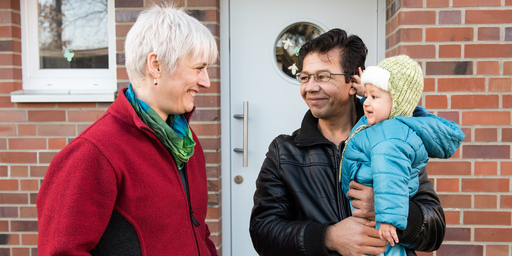 Eine blonde Frau spricht mit einem dunkelhäutigen Mann, der ein Kind auf den Armen hat. Sie stehen vor einem Hauseingang und lachen.