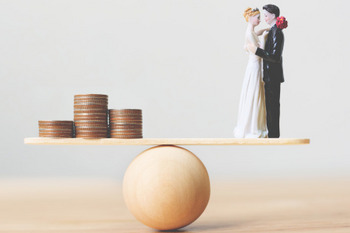 Eine Waage balanciert einen Stapel aus Kleingeld und eine Figur in Form eines Ehepaares auf einer Holzkugel.