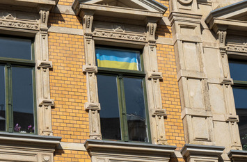 Der Blick trifft auf eine Hausfassade mit drei nah aneinander liegenden Fenstern. Im mittlerem Fenster hängt eine ukrainische Flagge.