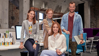 Vier junge Mitarbeiter:innen eines Start-ups sitzen beziehungsweise stehen in ihrem Büro in einem Fabrikloft vor einem Schreibtisch und lächeln in die Kamera.