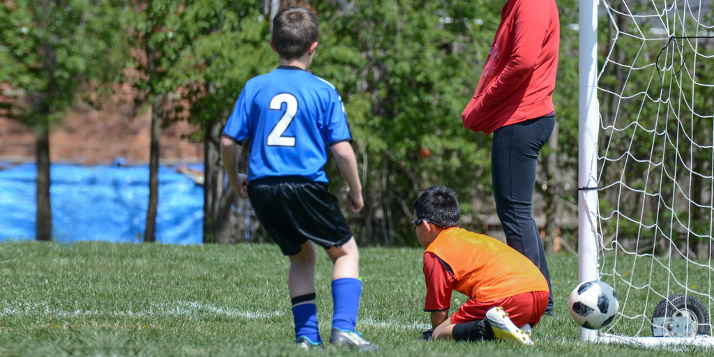 Ein Junge schießt bei einem Fußballspiel den Ball ins Tor. Der Torhüter des gegnerischen Teams, ebenfalls ein Junge, kniet am Boden und greift ins Leere.