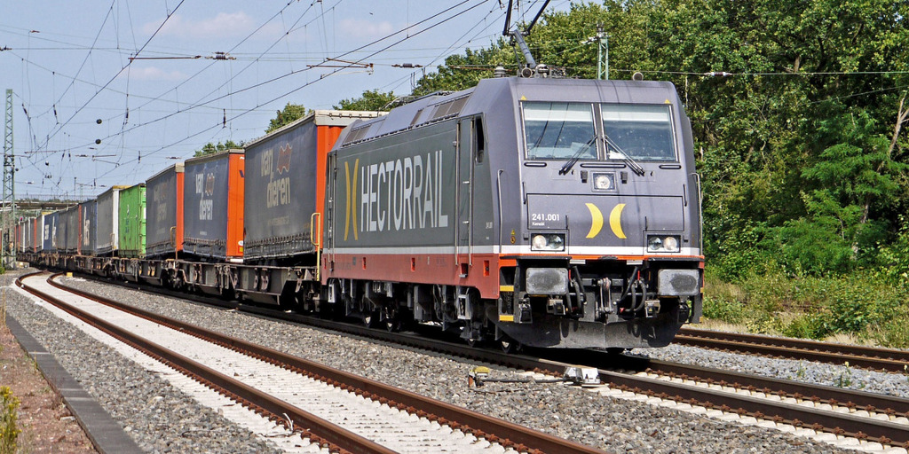 Eine Lokomotive mit mehreren Güterwaggons im Schlepptau fährt eine Bahnstrecke entlang. Über dem Zug hängen Oberleitungen. Im Hintergrund sind Bäume und eine Fußgängerbrücke zu sehen.