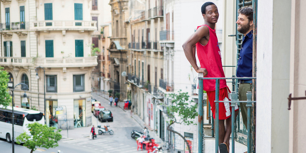 Zwei Männer stehen auf einem Balkon eines Hauses in der italienischen Stadt Palermo. Im Hintergrund Häuser und eine Straße, auf der Vespas fahren.