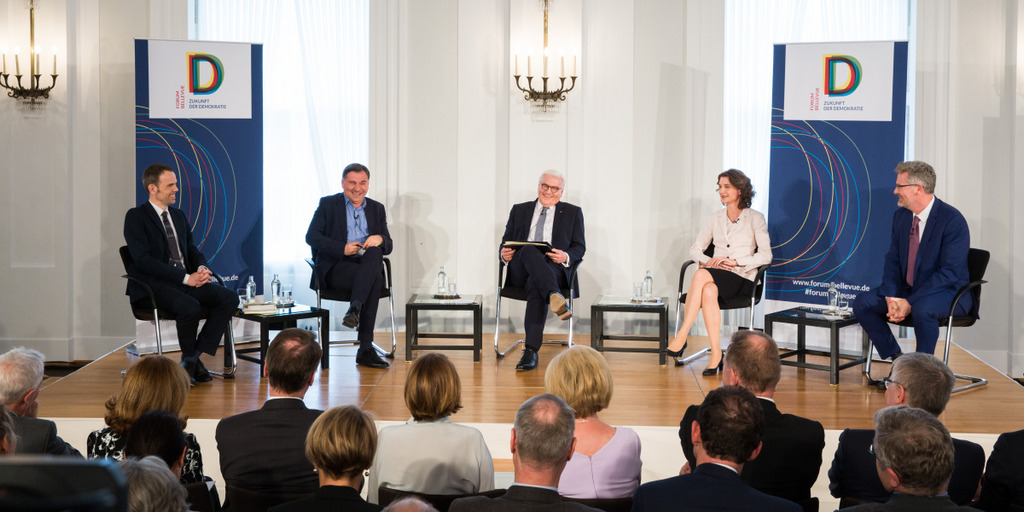 Blick aufs Podium beim Forum Bellevue, zu sehen ist der Bundespräsident im Kreis seiner Mitdiskutanten. Nach einer humorvollen Bemerkung lachen alle fünf Personen.