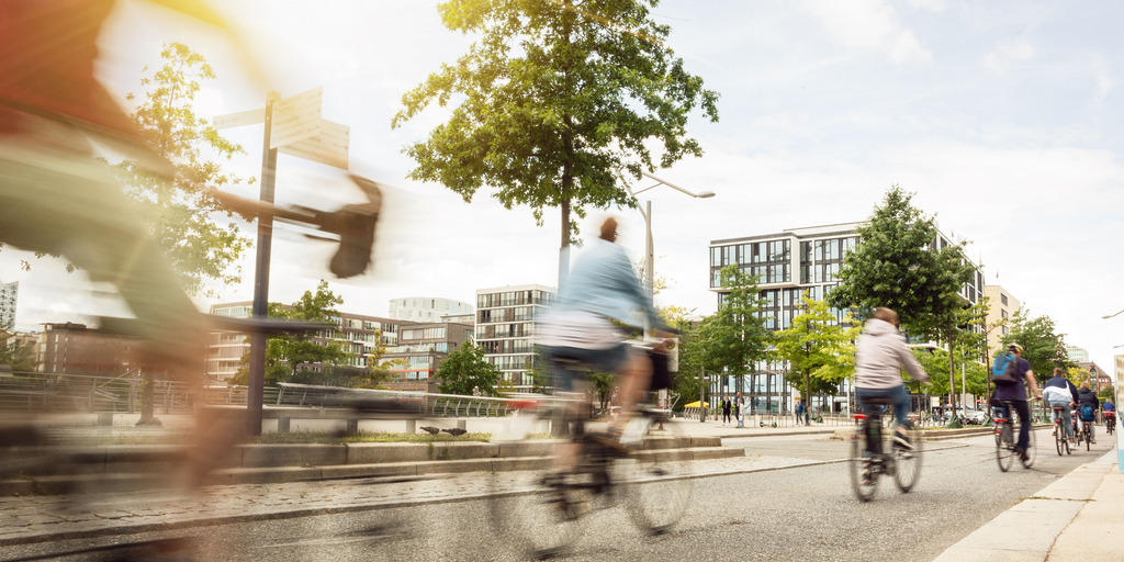 Auf dem Bild ist eine Fahrradstraße zu sehen. Die Fahrradfahren ist verschwommen dargestellt. Im Hintergrund stehen Bäume und Häuser. Es ist ein helles Bild.