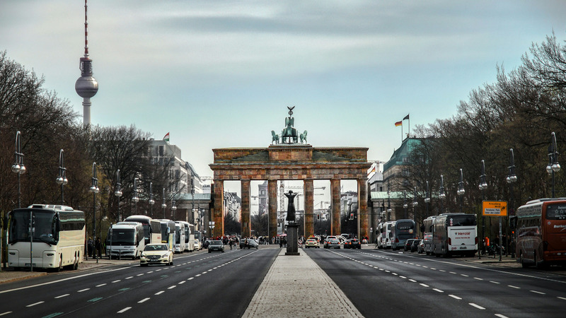 Das Bild zeigt das Brandenburger Tor in Berlin. Im Vordergrund verläuft eine breite Straße, flankiert von parkenden Bussen und Autos. Im Hintergrund ist der Berliner Fernsehturm zu sehen.