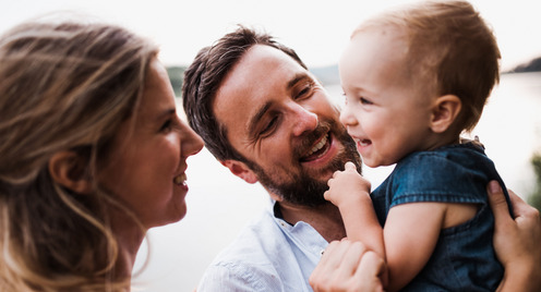 Ein junges Elternpaar mit ihrem kleinen Sohn. Der Vater hat den Sohn auf dem Arm, während die Mutter dem Jungen über den Rücken streichelt. Alle drei lächeln sich gegenseitig an.