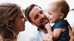Ein junges Elternpaar mit ihrem kleinen Sohn. Der Vater hat den Sohn auf dem Arm, während die Mutter dem Jungen über den Rücken streichelt. Alle drei lächeln sich gegenseitig an.