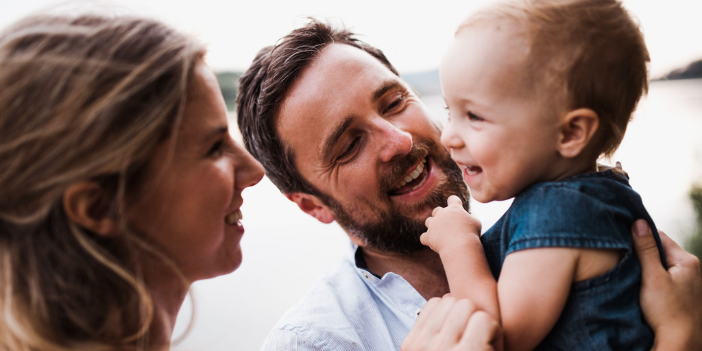 Ein junges Elternpaar mit ihrem kleinen Sohn. Der Vater hat den Sohn auf dem Arm, während die Mutter dem Jungen über den Rücken streichelt. Alle drei lächeln sich gegenseitig an.