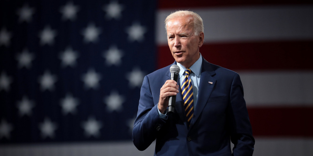 President-elect Joe Biden is giving a campaign speech in 2019 in Iowa. Behind him there is a giant US flag hanging on the wall.