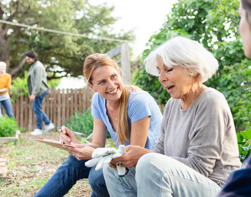 Zwei junge Frauen sitzen mit einer älteren Frau in einem Gemeinschaftsgarten und machen eine Pause von ihrer Gartenarbeit. Dabei unterhalten sie sich.