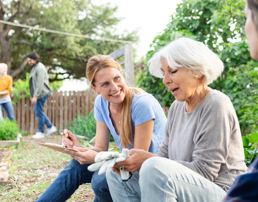 Zwei junge Frauen sitzen mit einer älteren Frau in einem Gemeinschaftsgarten und machen eine Pause von ihrer Gartenarbeit. Dabei unterhalten sie sich.