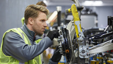 Blonder junger Mann ist von der Seite zu sehen, wie er auf Augenhöhe an einem Motor mit einem Schraubenzieher schraubt. Er träft eine neongelbe Warnweste und einen grauen Arbeitsanzug.(© <p>© Getty Images/iStockphoto/Morsa Images</p>)