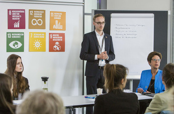 Auf dem Foto ist eine Präsentationssituation zu sehen. Drei Referent:innen stehen und sitzen vor einem Publikum.