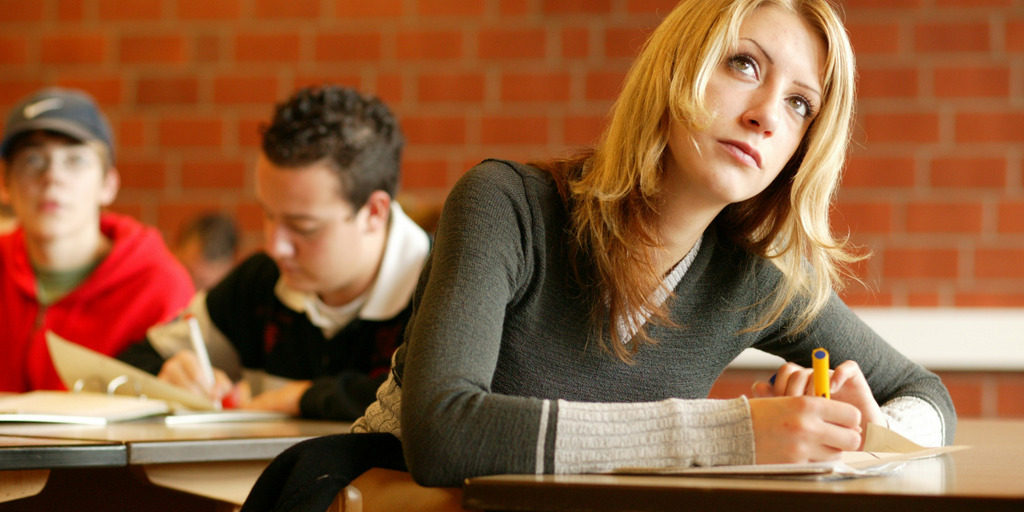 Eine junge Frau sitzt konzentriert im Schulunterricht, hält einen Füller in der rechten Hand und liest etwas von der Tafel ab. Im Hintergrund sieht man zwei ihrer Mitschüler.