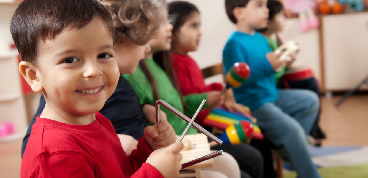 (© @ fatihhoca / iStockphoto) Kinder sitzen im Kreis und musizieren mit Triangel, Rassel und Trommel