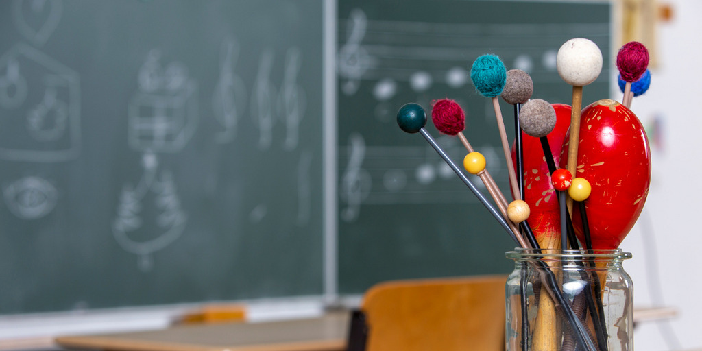 Fotoaufnahme eines Musik-Klassenzimmers. Im Vordergrund steht ein durchsichtiges Glasgefäß mit Schlägeln und Maracas. Im Hintergrund ist eine Tafel zu sehen.