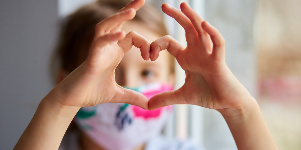 Ein Mädchen, das einen Mundschutz trägt, formt mit seinen Fingern ein Herzsymbol und streckt es der Kamera entgegen.