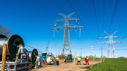 Das Bild zeigt eine Baustelle für Hochspannungsleitungen in einer ländlichen Umgebung. Mehrere Arbeiter in Sicherheitskleidung und Helmen überwachen die Installation oder Wartung von Stromleitungen. Große Kabeltrommeln und Maschinen sind im Vordergrund zu sehen, während sich ein hoher Strommast im Zentrum des Bildes befindet. Im Hintergrund erstrecken sich weitere Hochspannungsleitungen und Masten unter einem klaren blauen Himmel.