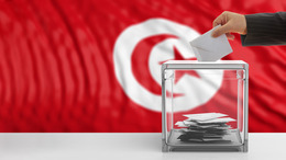 Hand wirft einen Stimmzettel in Wahlurne vor tunesischer Fahne