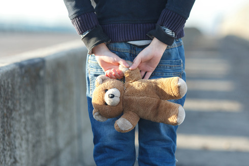 Kind mit einem Teddy in den hinter dem Rücken verschränkten Armen