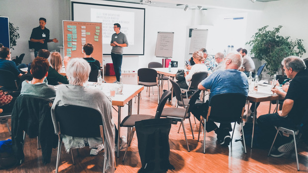 Open-Data-Workshop in Münster. Etwas 25 Personen sitzen in einem Raum. 2 Moderaoren stehen vorne an einer Metaplanwand.