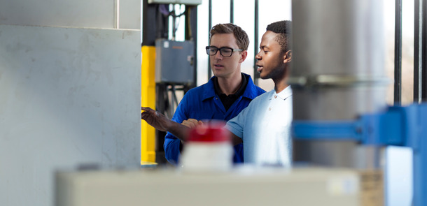Ein hellhäutiger Ingenieur erklärt einem dunkelhäutigen Auszubildenden die Bedienung einer Maschine in einer Fabrik.