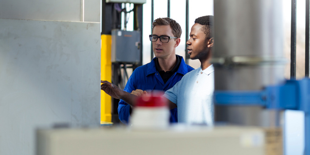 In a factory, a white engineer explains to a black apprentice how to user a specific machine.