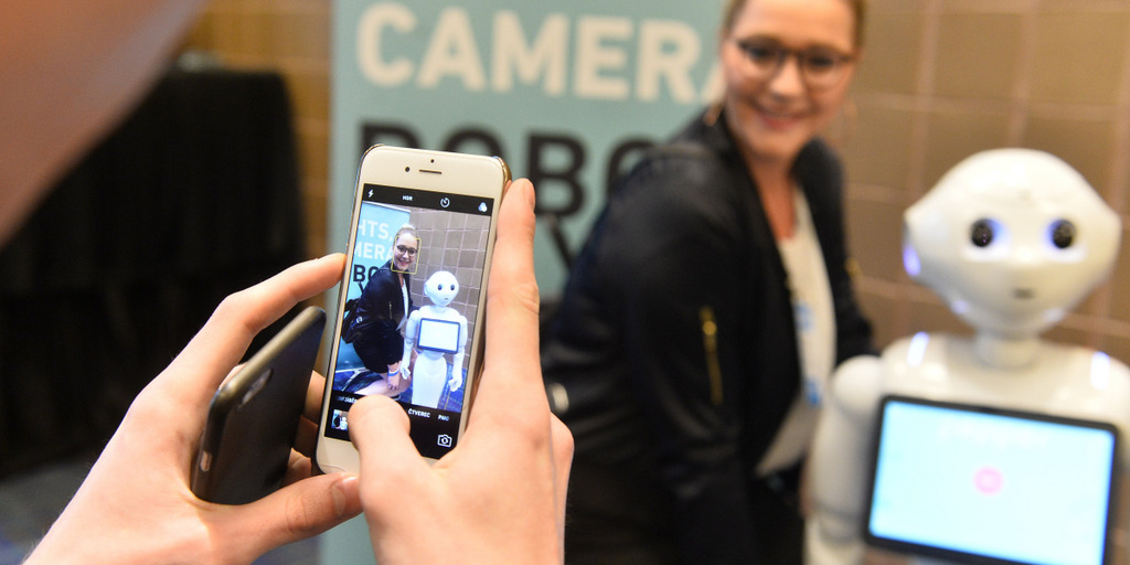 Eine Hand hält ein Smartphone in der Hand und fotografiert eine Frau und einen Roboter.