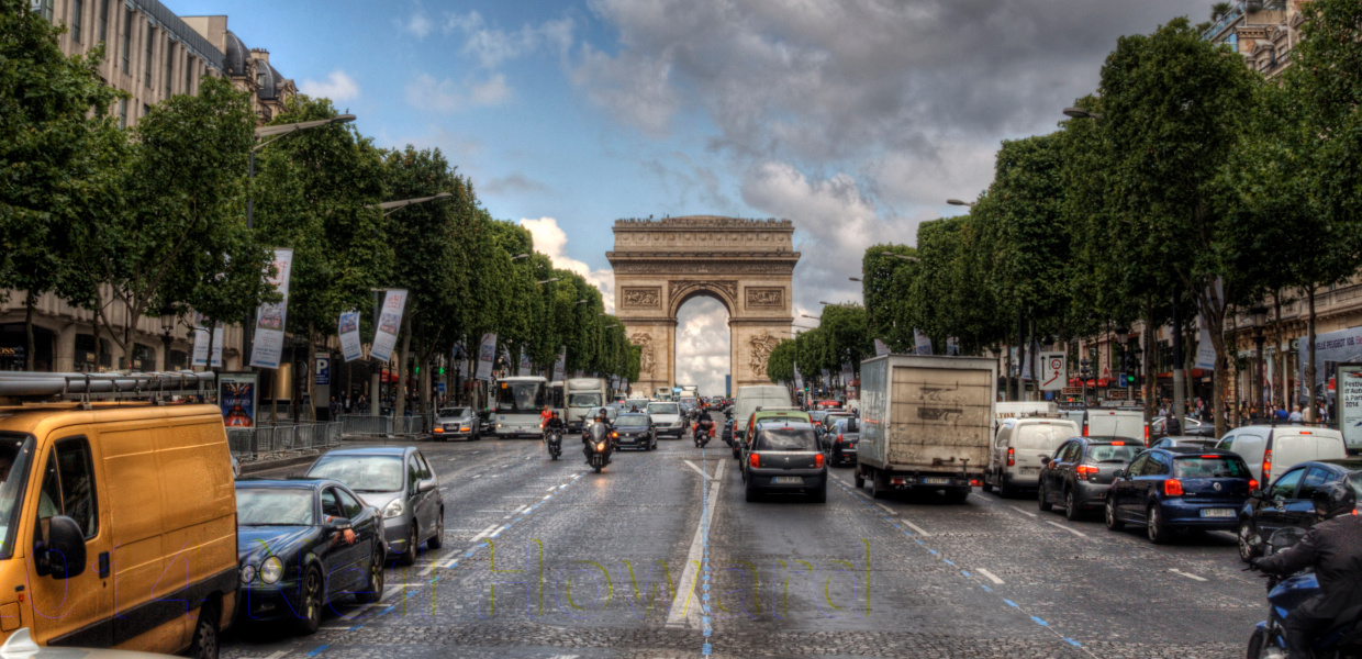 Blick auf den Champs Elysees in Paris. Im Hintergrund der Triumphbogen. Davor die Straße, befahren von Autos, LKWs und Mopeds. Die Straße ist gesäumt von Bäumen. Dahinter rechts und links Cafes, Restaurants, Sonnenschirme und Flaggen.