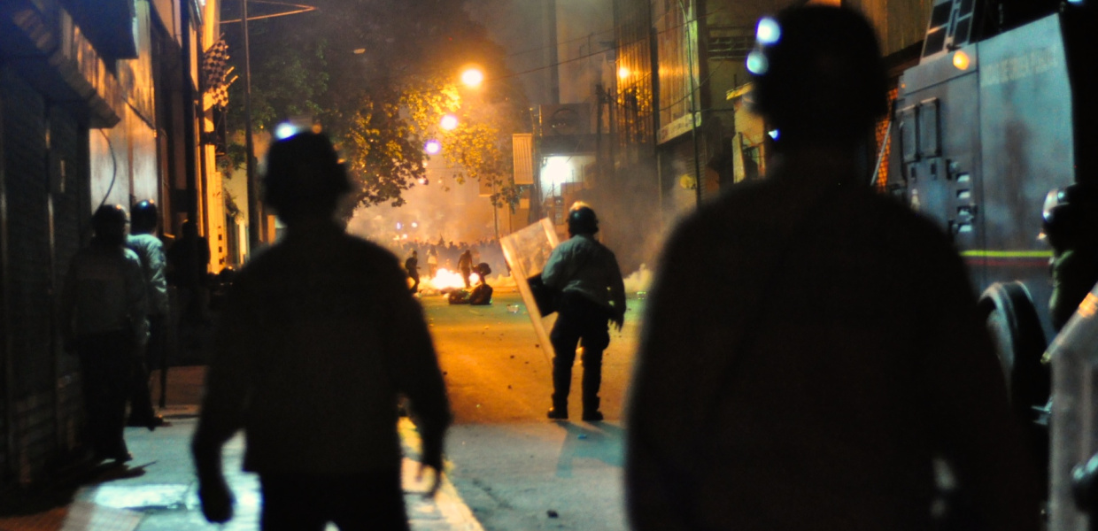 Proteste gegen die Regierung von Staatspräsident Nicolás Maduro in Venezuela: Auf einer Straße in der Hauptstadt Caracas haben sich am 15. Februar 2014 Polizisten mit Helmen, Schilden und einem gepanzerten Fahrzeug postiert und blicken auf die in der Ferne demonstrierende Menschenmenge. Zwischen ihnen und den Demonstranten brennt ein Feuer auf der Straße, das einzelne Demonstranten weiter anfachen. Titelbild für die Onlinemeldung und Pressemitteilung zum BTI 2018.