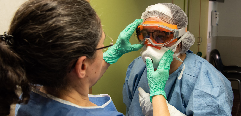 Eine Krankenpflegerin prüft bei einer anderen Krankenpflegerin den richtigen Sitz ihrer Atemschutzmaske und Schutzbrille gegen das Coronavirus