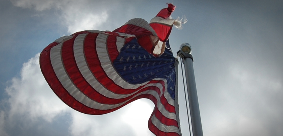 Against the background of an eerily dark grey sky, a slightly tattered US flag is flying on a flagpole.