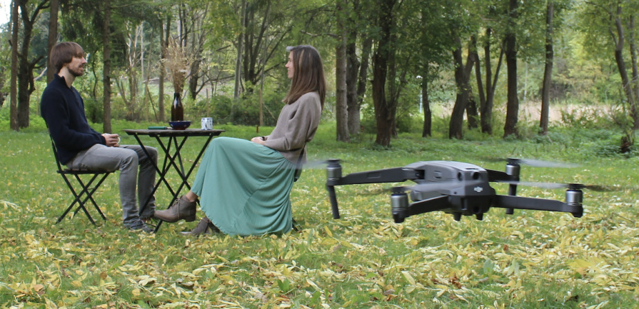 Mann und Frau sitzen im Wald gemeinsam an einem Tisch. Vor ihnen fliegt eine Drohne.