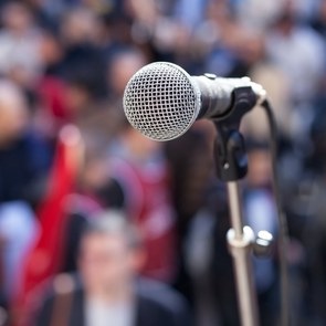 Ein Mikrofon auf einem Ständer. Dahinter unscharf zu sehen eine Menschenmenge mit Fahnen.