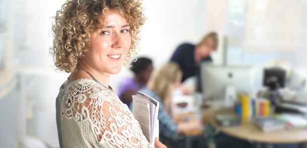 Frau mit Broschüren im Arm lächelt in die Kamera. Im Hintergrund sind schemenhaft ein Arbeitsplatz und 3 weitere Personen zu erkennen.