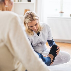 Eine junge Pflegekarft zieht einer älteren Frau zu Hause Pantoffeln an.