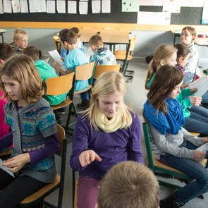 viele Kinder sitzen sich auf ihren Stühlen im Klassenraum gegenüber, sie halten ein Blatt Papier in der Hand