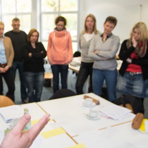 Lehrer und Lehrerinnen stehen in einem Klassenraum und hören dem Redner zu