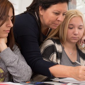 Eine Lehrerin beugt sich von hinten zwischen zwei Schülerinnen durch und korrigiert etwas im Heft der Schülerin
