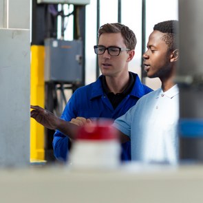 Ein hellhäutiger Ingenieur erklärt einem dunkelhäutigen Auszubildenden die Bedienung einer Maschine in einer Fabrik.