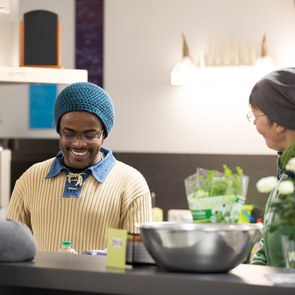 Ein dunkelhäutiger Geflüchteter kocht im Allerlüd, einem Haus für Austausch und Begegnung in Lüchow (Niedersachsen), gemeinsam mit einer Helferin in der Gemeinschaftsküche. Beide lachen.